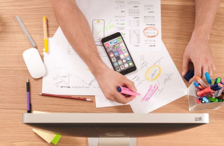 Persona diseñando wireframes de aplicación móvil en escritorio con iPhone, marcadores y computadora